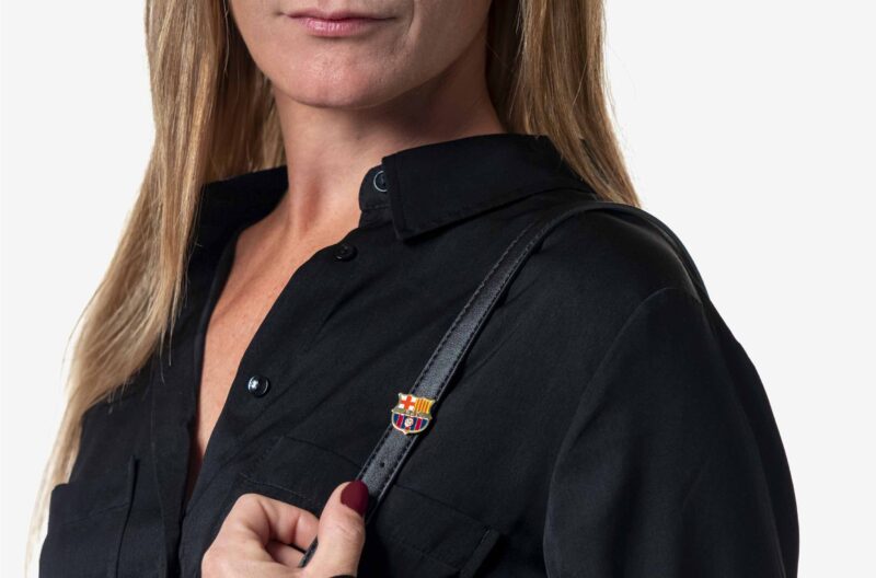 Modelo femenina de pelo rubio y piel blanca con una camisa negra vistiendo en la correa de su bolso, la insignia del FC Barcelona acabada en oro de 18K y con un diamante engastado en su centro.