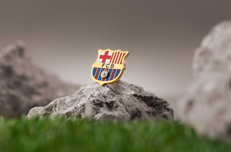 La emblemática insignia del escudo del FC Barcelona, presentada sobre una piedra en forma triangular rodeada de césped y otras piedras. La brillante insignia, bañada en oro y esmalte, resalta, reflejando la grandeza del club